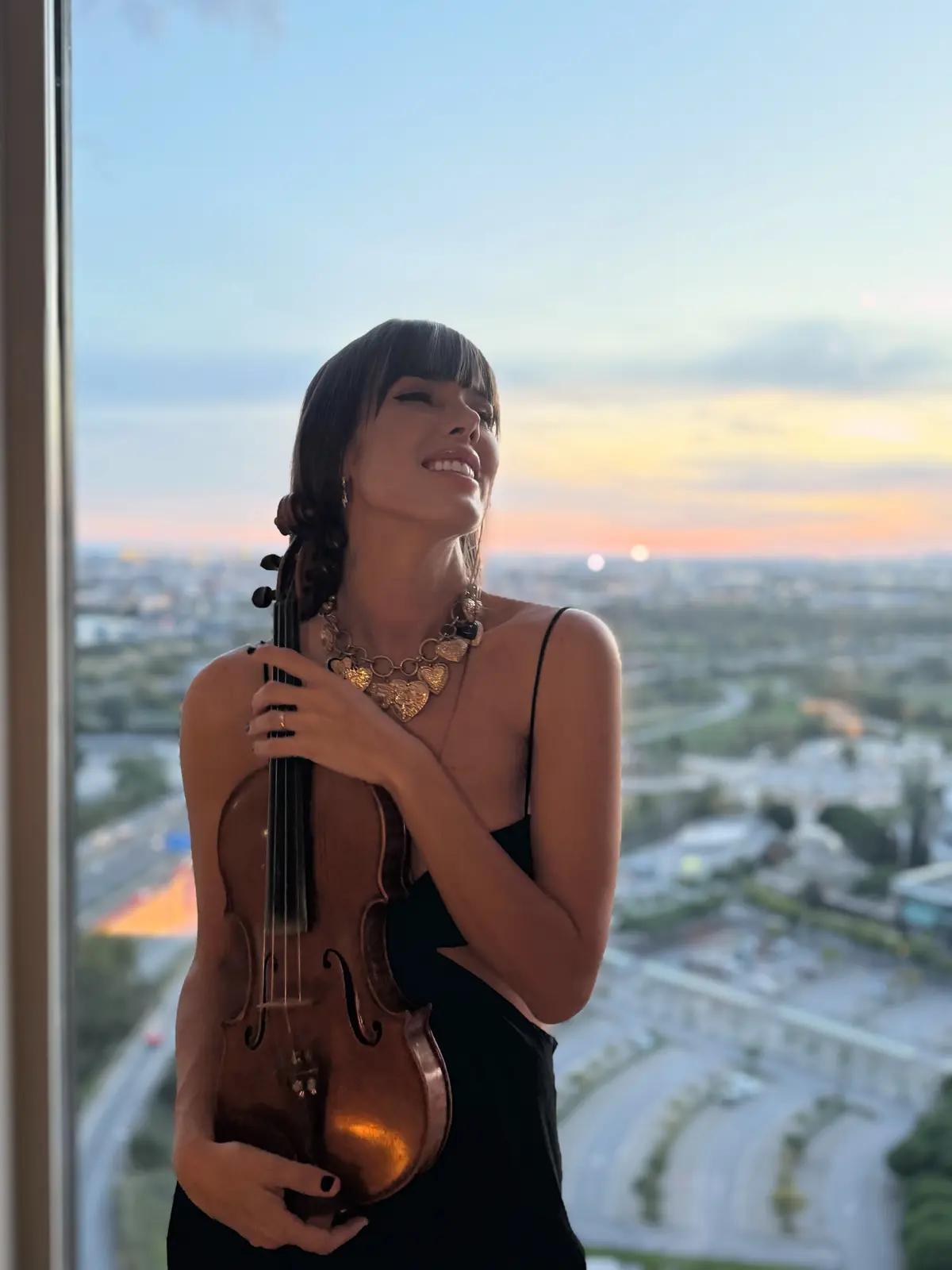 Astrid Torrente sonriendo con su violín acústico al atardecer frente a una vista panorámica