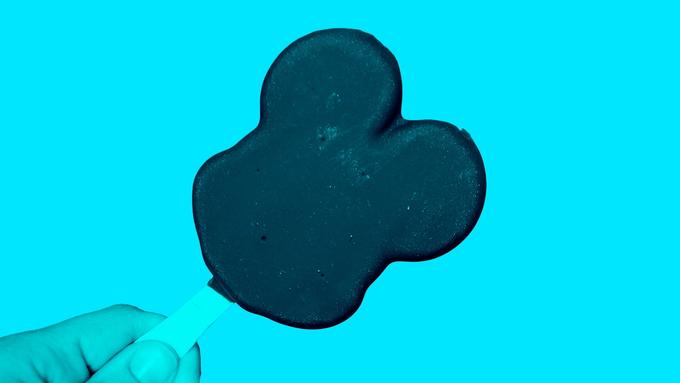 A hand holding a Mickey Mouse-shaped ice cream bar against a teal background