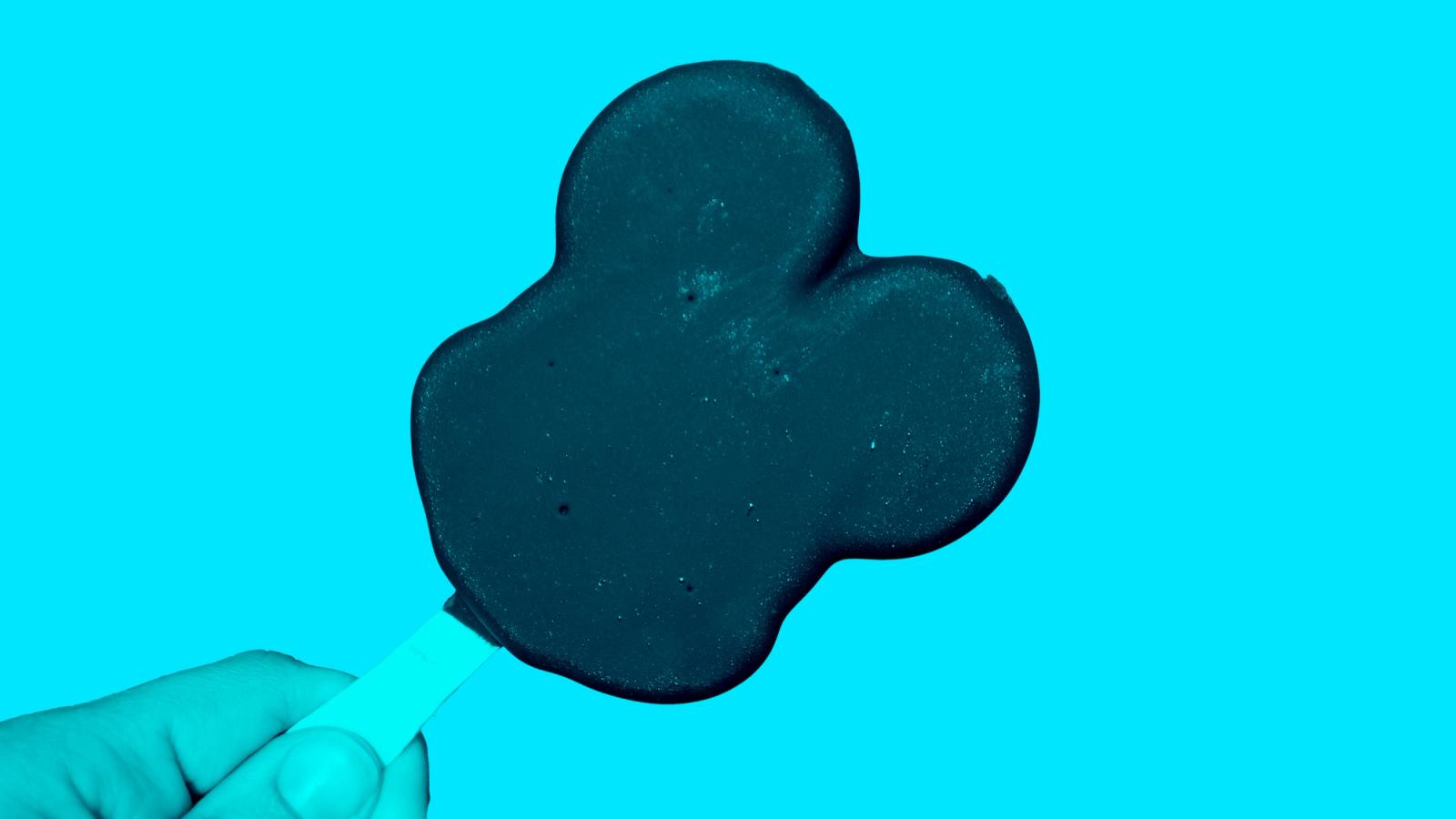 A hand holding a Mickey Mouse-shaped ice cream bar against a teal background