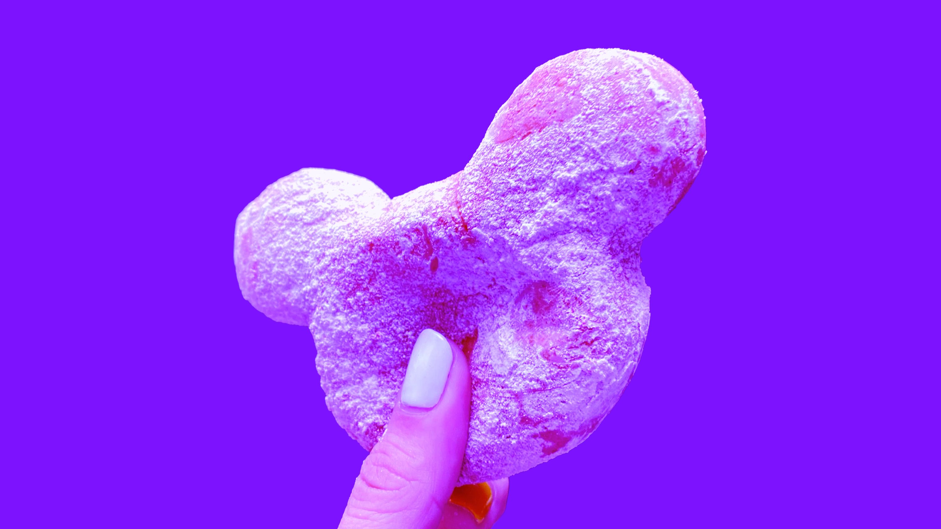 A hand holding a Mickey-shaped beignet dusted with powdered sugar, on a purple background