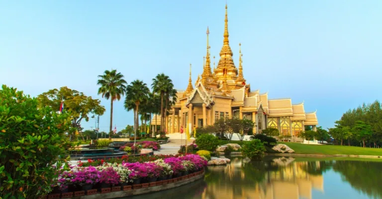 A golden Thai temple beside a lake, framed by palm trees and flowering gardens