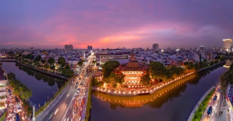 Aerial view of Ho Chi Minh City at dusk, with traffic streaming past a lit-up temple on the canal