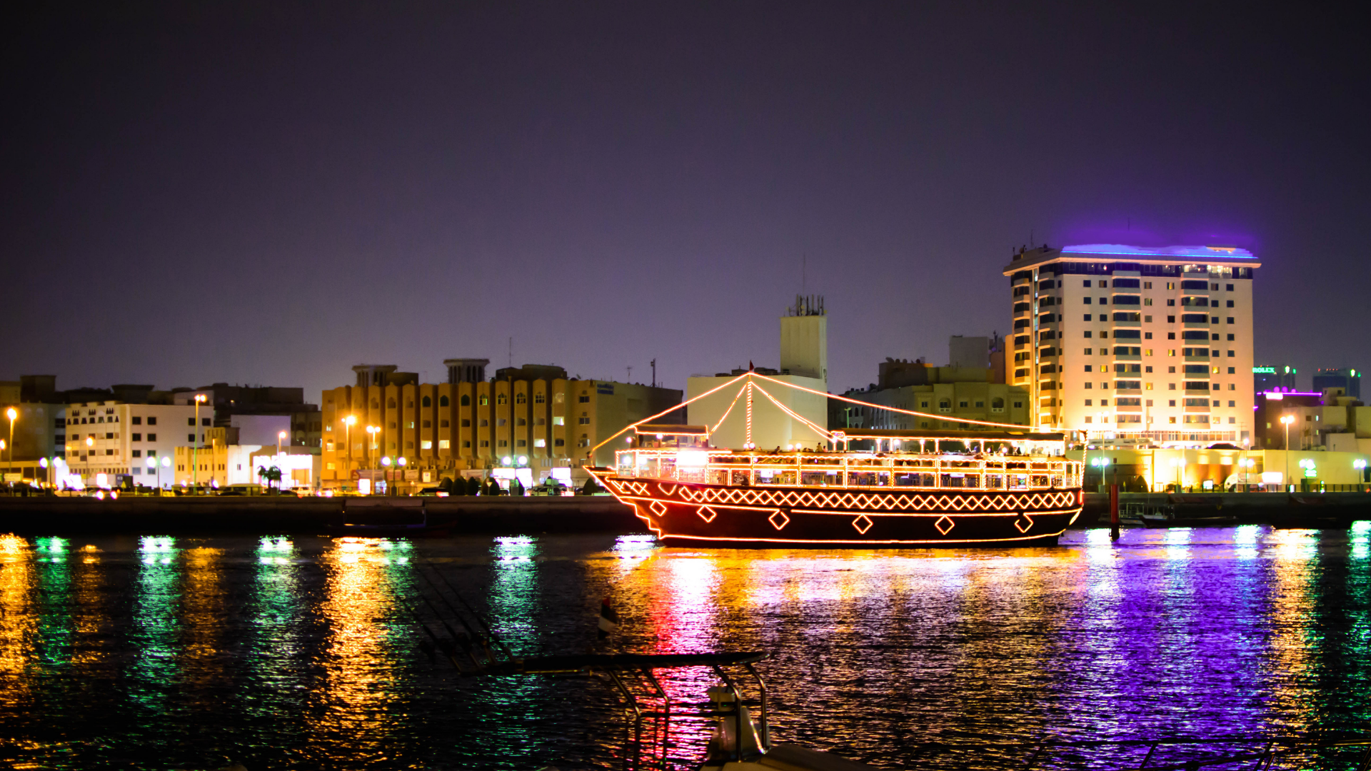 An illuminated dhow boat sails along a city waterway at night, with lit-up buildings in the background and colorful reflections on the water