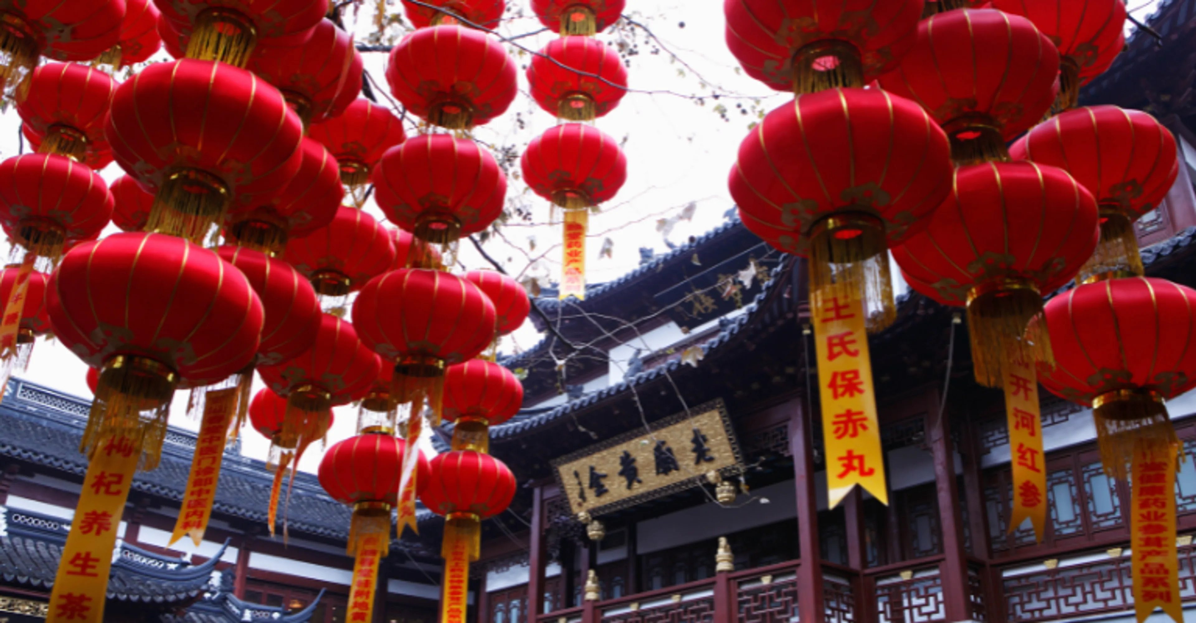 Rows of red Chinese lanterns with gold trim hang beneath a traditional Chinese building's eaves, with vertical red and gold banners bearing Chinese characters hanging among them