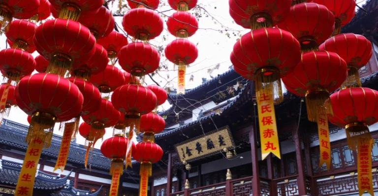 Rows of red Chinese lanterns with gold trim hang beneath a traditional Chinese building's eaves, with vertical red and gold banners bearing Chinese characters hanging among them
