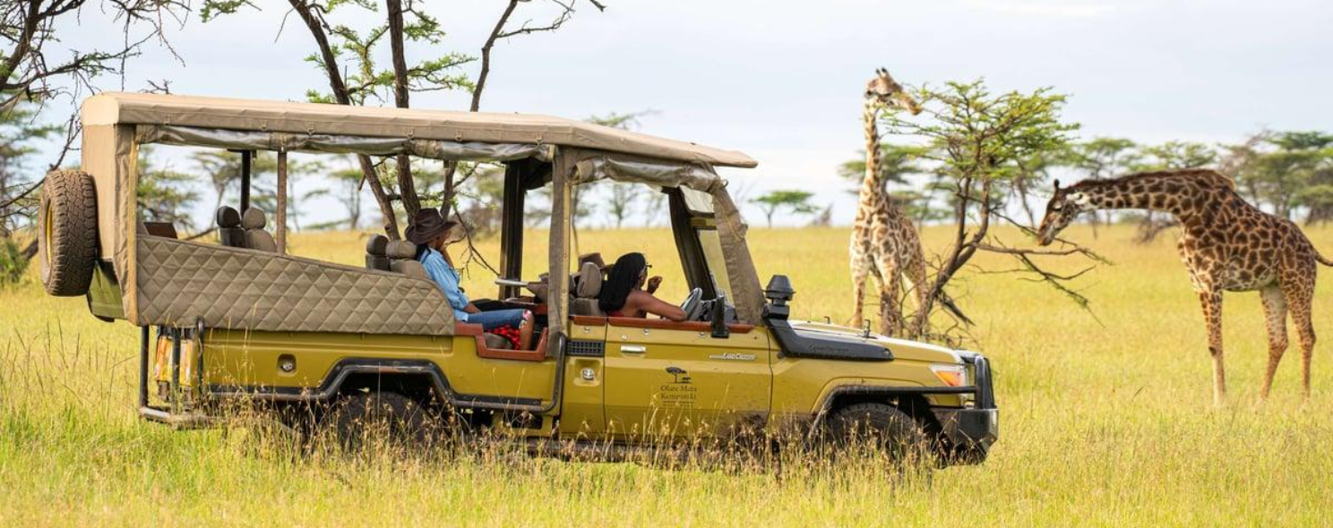 A yellow open-sided safari vehicle with tourists drives past two giraffes standing among acacia trees on the savanna.