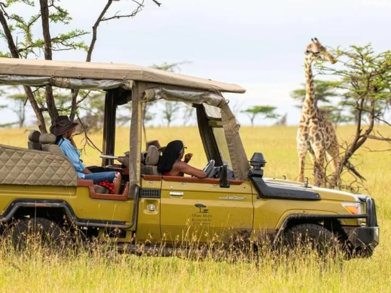 A yellow open-sided safari vehicle with tourists drives past two giraffes standing among acacia trees on the savanna.