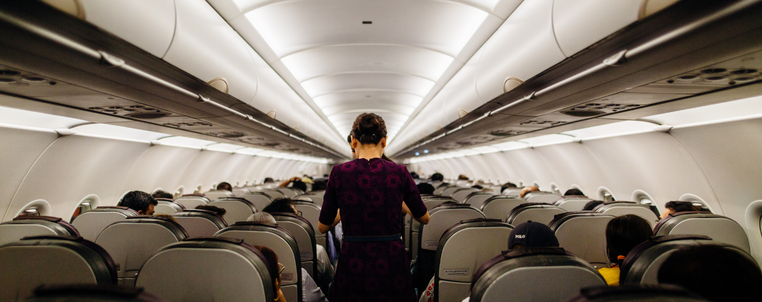 View down the aisle of an airplane cabin from the rear, showing a flight attendant in a purple patterned dress walking toward the front past rows of seated passengers
