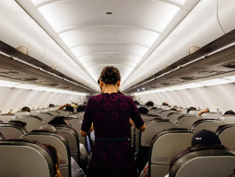 View down the aisle of an airplane cabin from the rear, showing a flight attendant in a purple patterned dress walking toward the front past rows of seated passengers