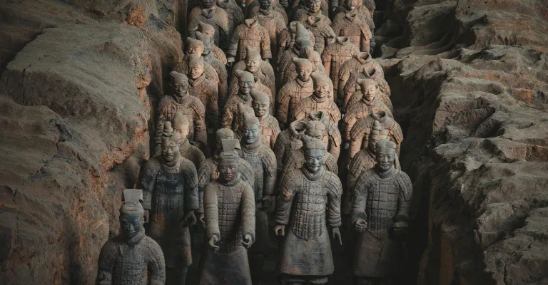 Rows of ancient terracotta warrior statues with detailed armor and hairstyles stand in an excavated pit with rough earthen walls, part of the Terracotta Army