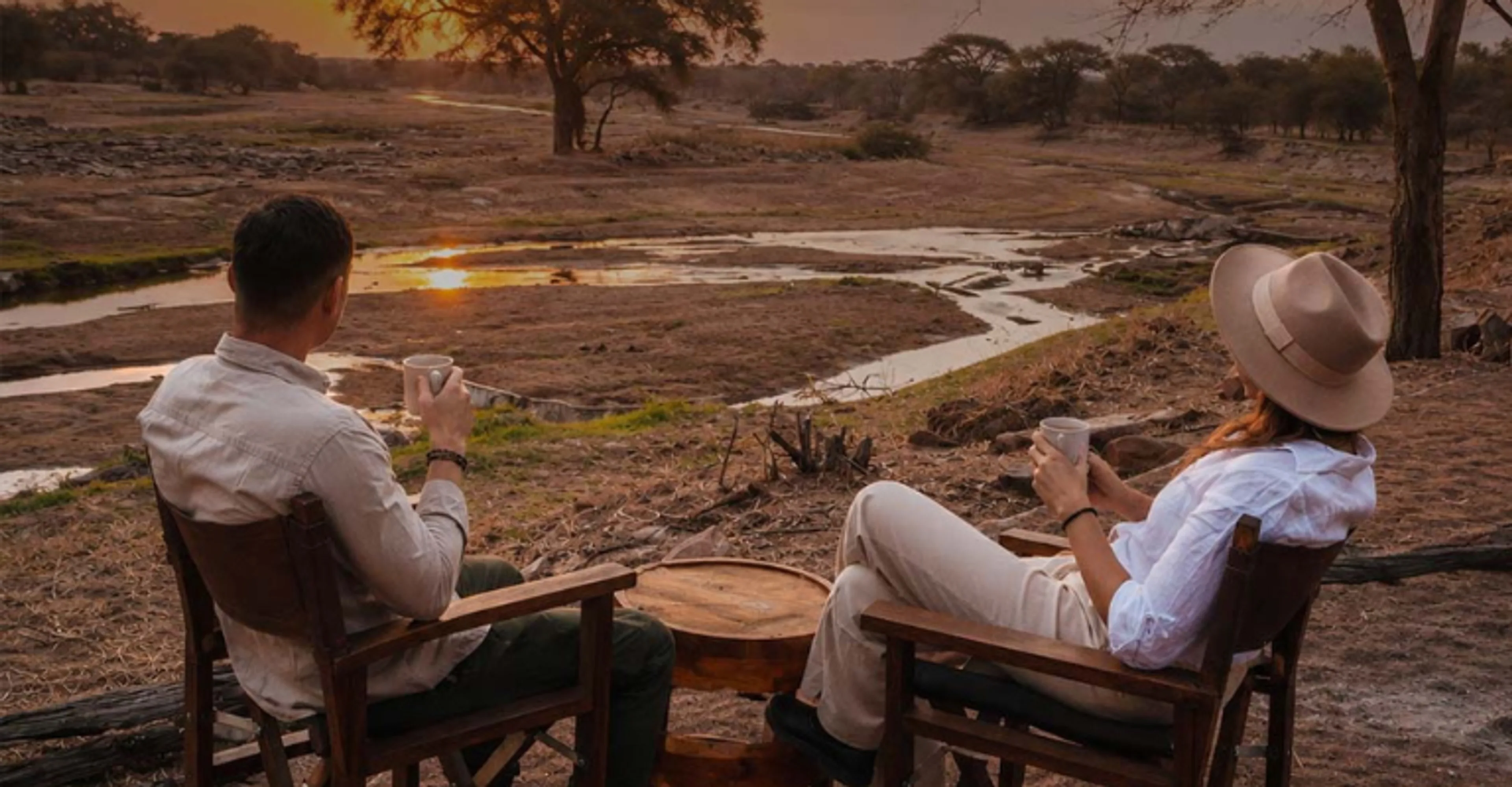 A couple sits with warm drinks overlooking a watering hole at dusk, watching the sunset reflect across the water from camp chairs