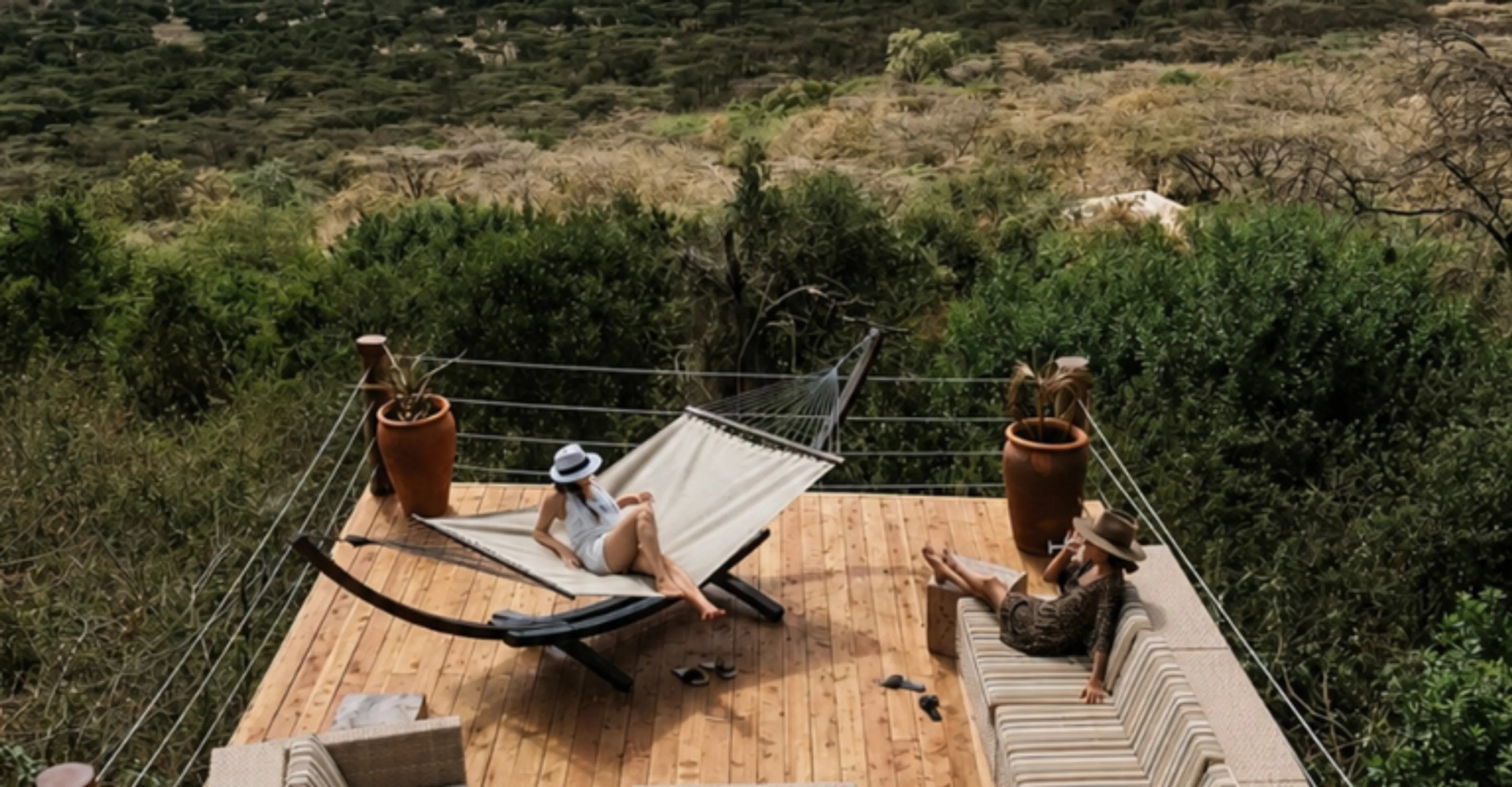 An aerial view of a wooden lodge deck with a woman relaxing in a hammock and another woman seated on a cushioned bench, overlooking dense green bushland.