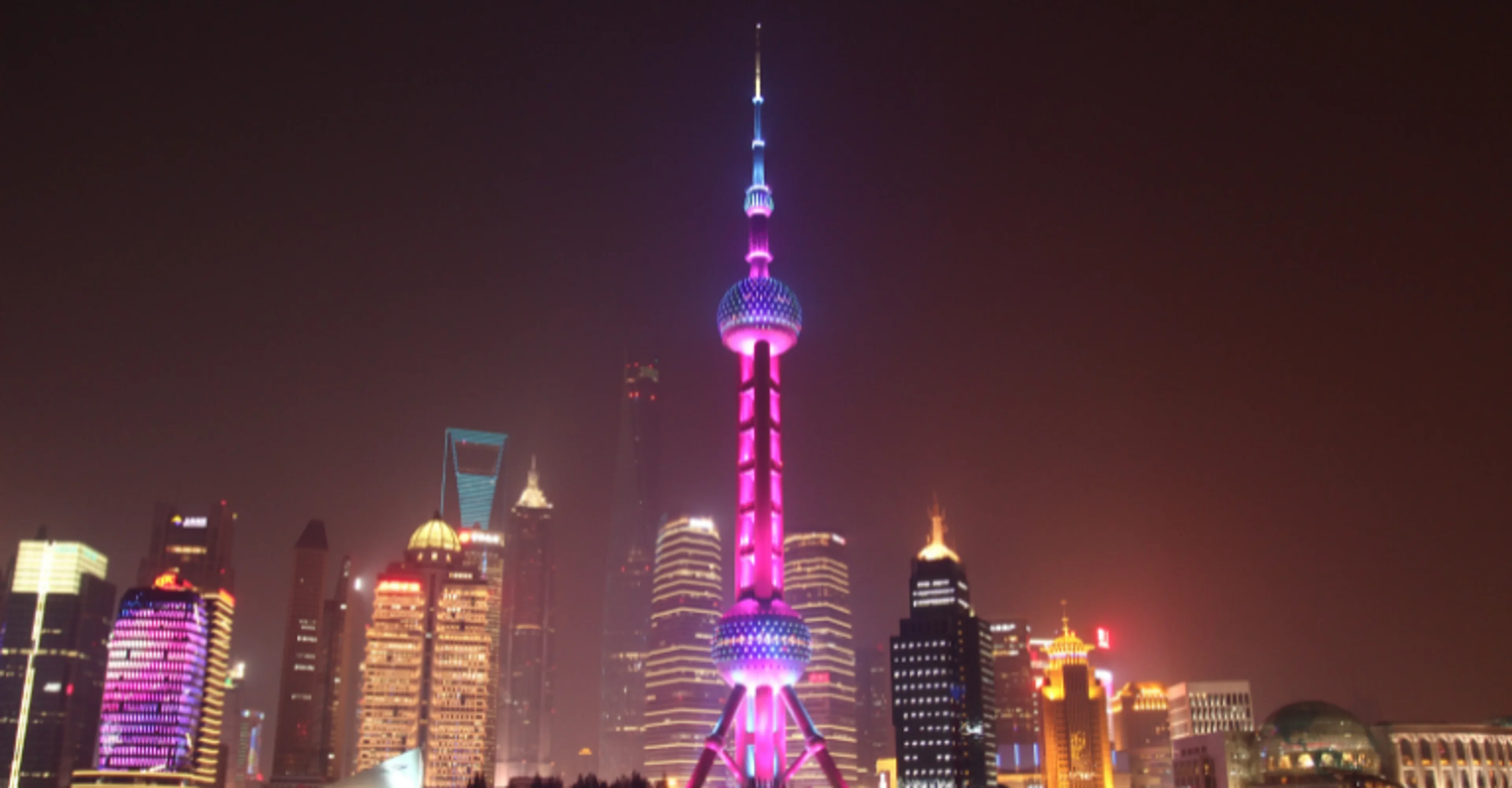The Oriental Pearl Tower illuminated in pink and purple lights against Shanghai's night skyline, surrounded by other lit skyscrapers under a hazy dark sky.
