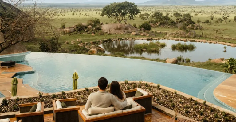 A couple sits on an outdoor lounge sofa overlooking an infinity pool that blends into a watering hole, with a grassy savanna, scattered trees, and distant hills in the background.