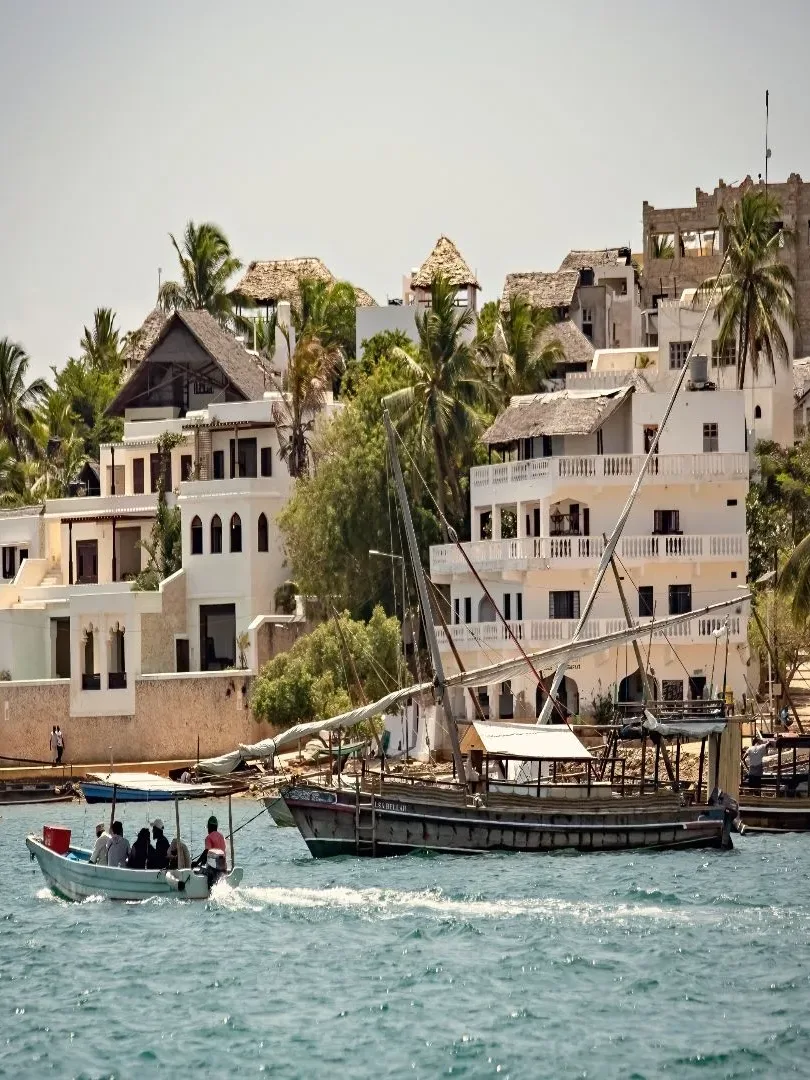 Traditional dhow boats sailing along the historic coastline of Lamu Island with coral-stone buildings and palm trees at sunset