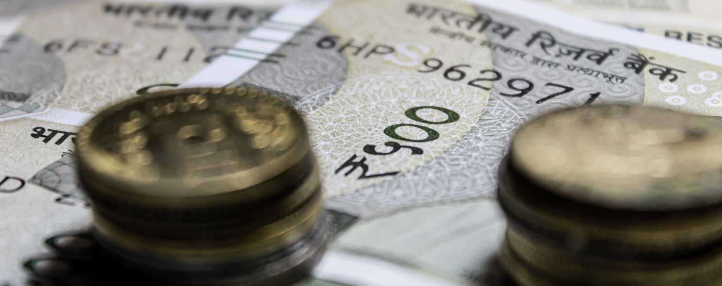 Close-up of stacked Indian coins in front of Indian Reserve Bank 500 rupee banknotes, with denomination numerals and serial numbers visible on the currency.