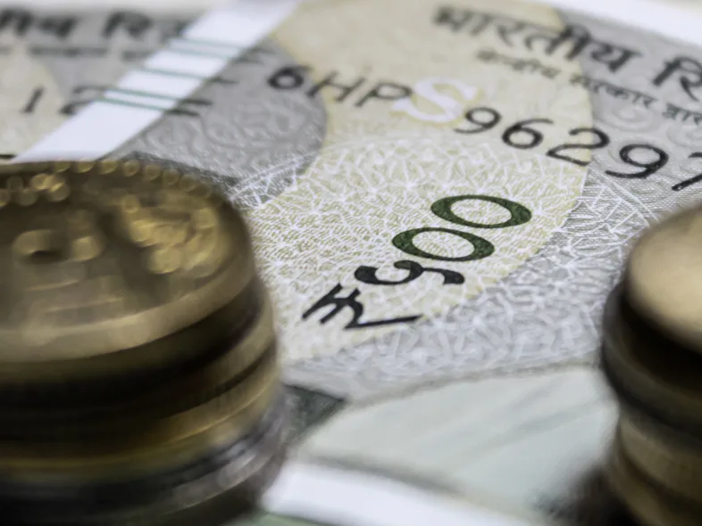 Close-up of stacked Indian coins in front of Indian Reserve Bank 500 rupee banknotes, with denomination numerals and serial numbers visible on the currency.