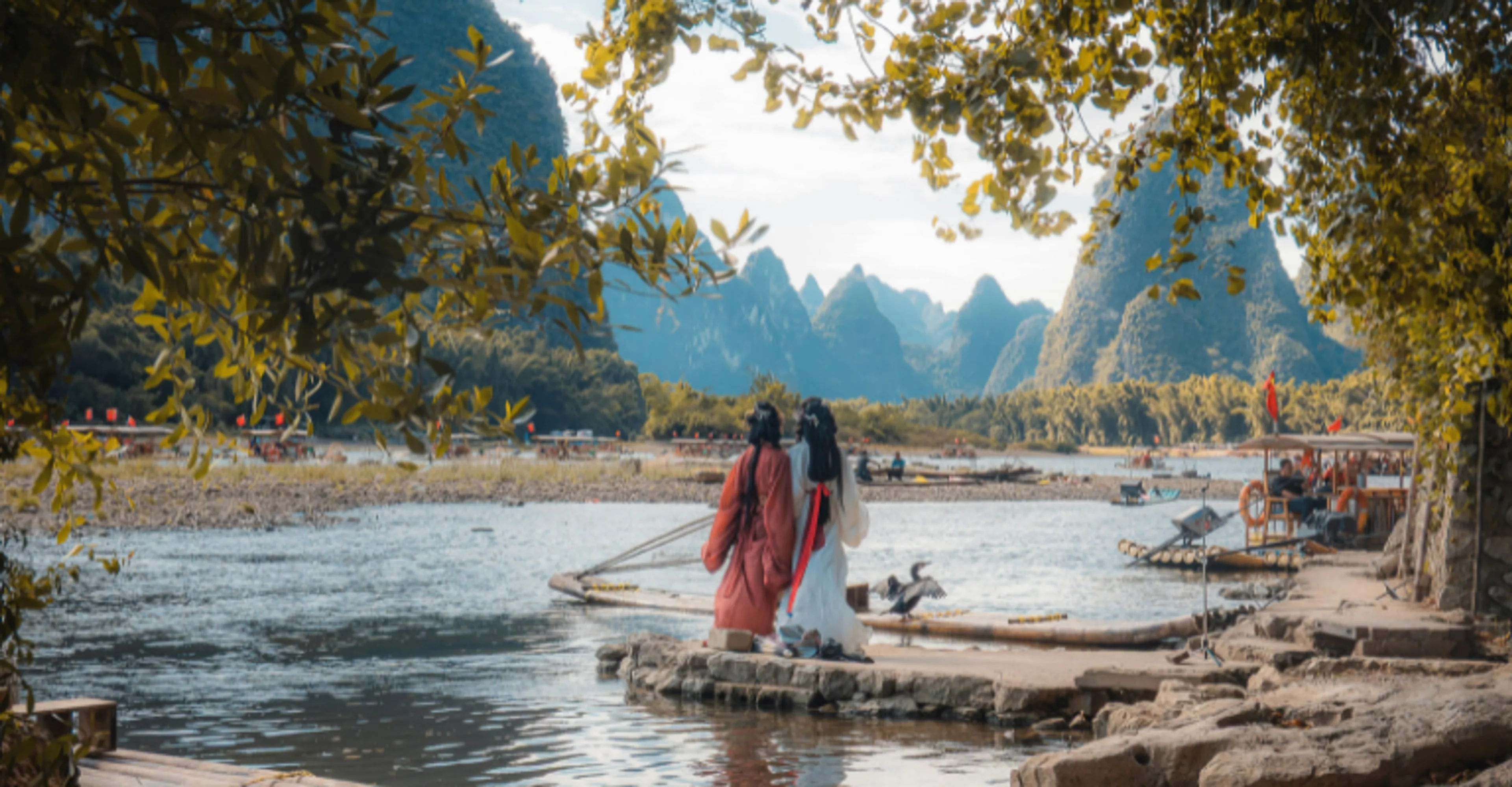 Two people in traditional red and white robes stand on a rocky riverbank framed by hanging tree branches, looking out at dramatic karst limestone peaks along the Li River, with bamboo rafts and boats on the water.