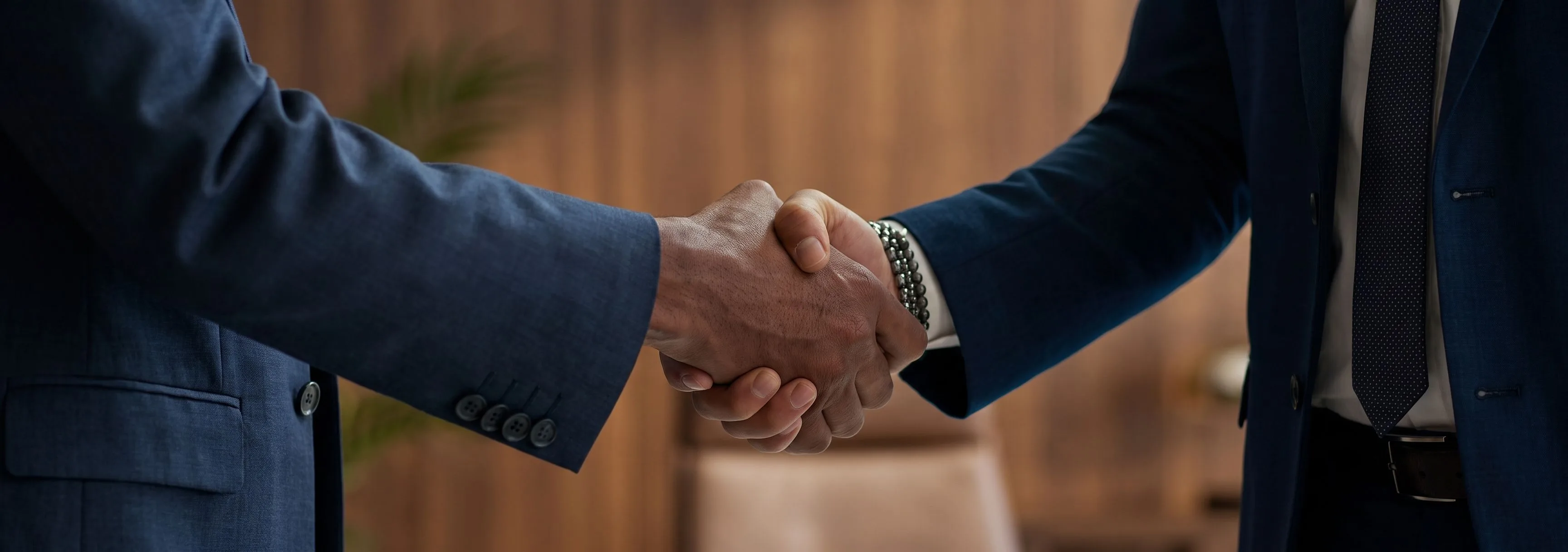 Two people in navy business suits shaking hands in an office with warm wood-paneled walls.