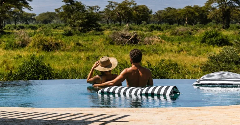 A couple relaxes in an infinity pool at the edge of a lush green safari landscape, resting on a striped pool float with bushveld stretching into the distance.