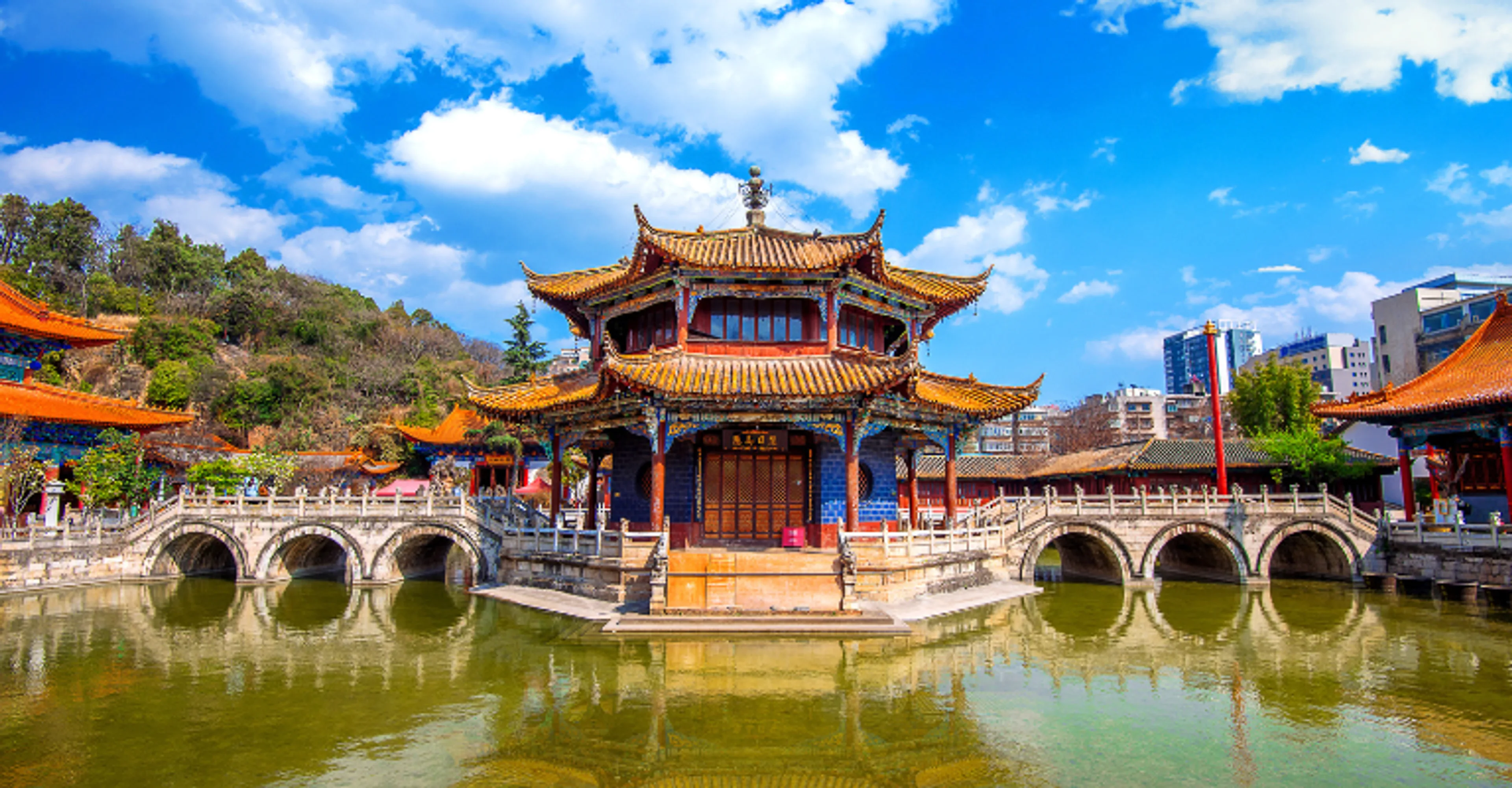 A vividly painted red and gold Chinese temple pavilion with multi-tiered curved roofs stands on an island surrounded by a green pond, connected by arched stone bridges, with city buildings visible in the background under a bright blue sky