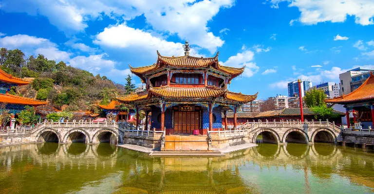 A vividly painted red and gold Chinese temple pavilion with multi-tiered curved roofs stands on an island surrounded by a green pond, connected by arched stone bridges, with city buildings visible in the background under a bright blue sky