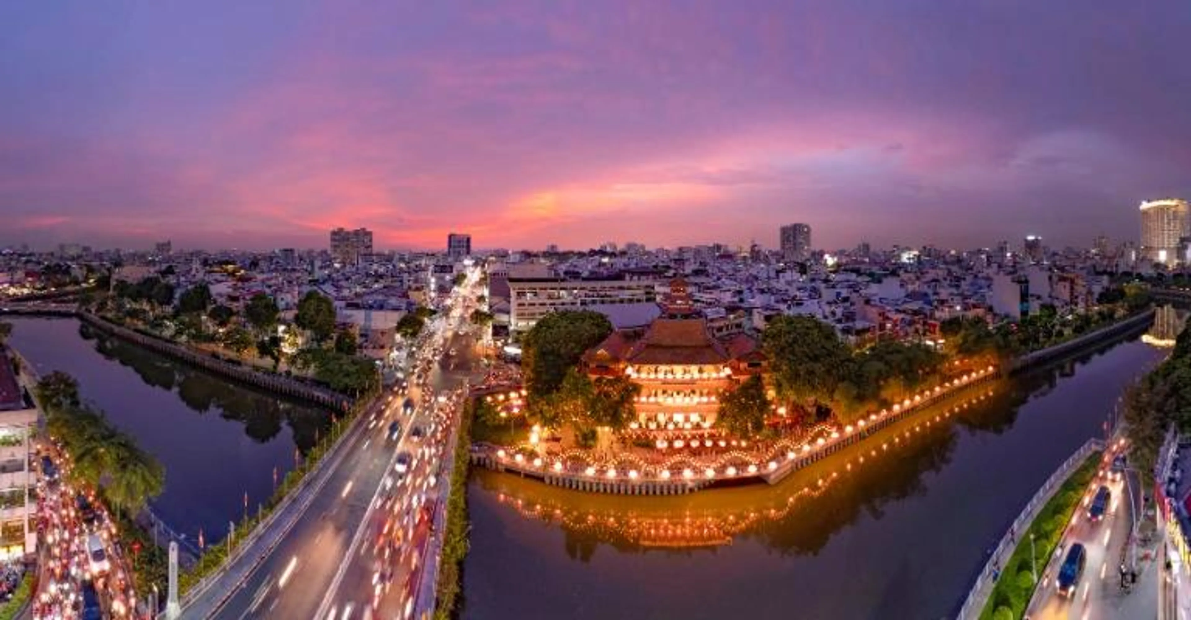 Aerial view of Ho Chi Minh City at dusk, with traffic streaming past a lit-up temple on the canal