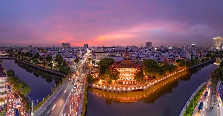 Aerial view of Ho Chi Minh City at dusk, with traffic streaming past a lit-up temple on the canal