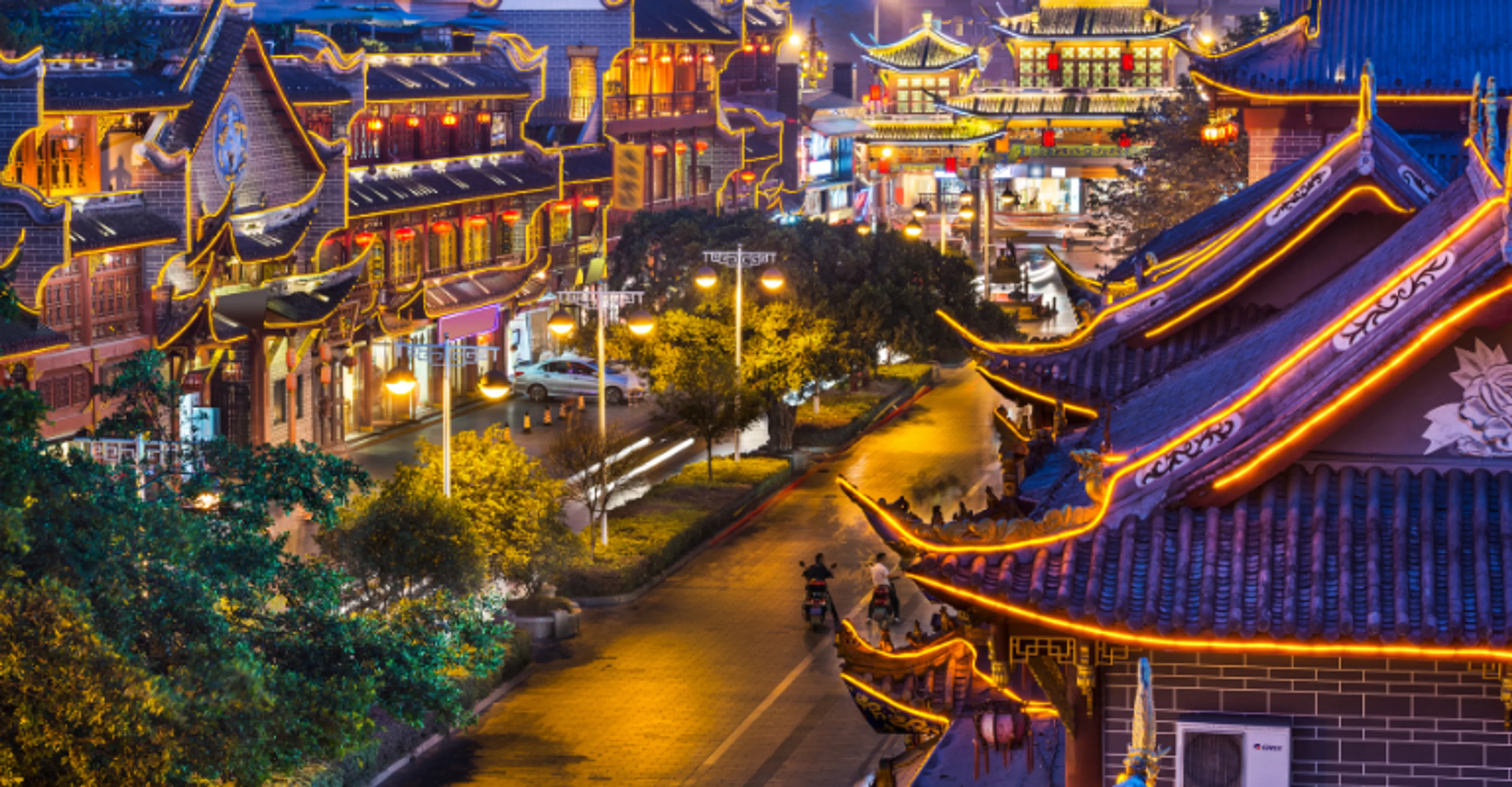 An elevated night view of a traditional Chinese street lined with buildings outlined in warm golden lights, featuring upturned pagoda-style roofs, red lanterns, and a road with a few motorbikes passing through
