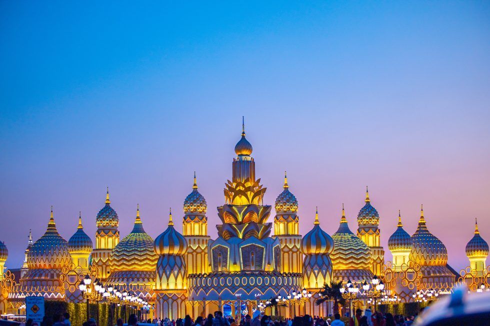 Ornate golden domed structures with intricate patterns illuminated at dusk, with a gradient sky and street lamps in front.