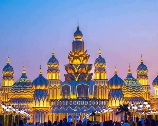 Ornate golden domed structures with intricate patterns illuminated at dusk, with a gradient sky and street lamps in front.