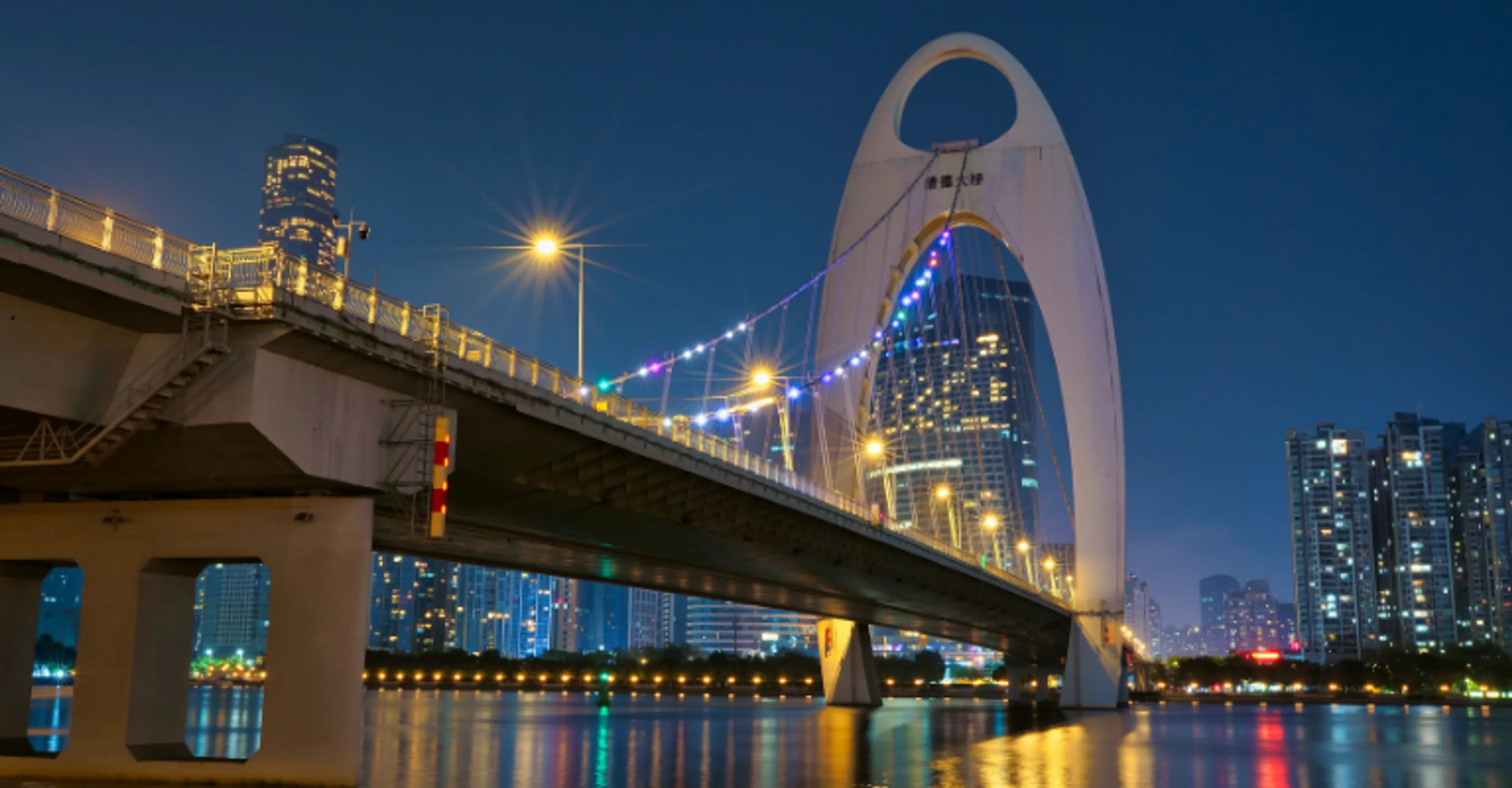 A large arched suspension bridge decorated with blue lights spans a river at night, with illuminated high-rise buildings lining the waterfront and reflecting in the water below