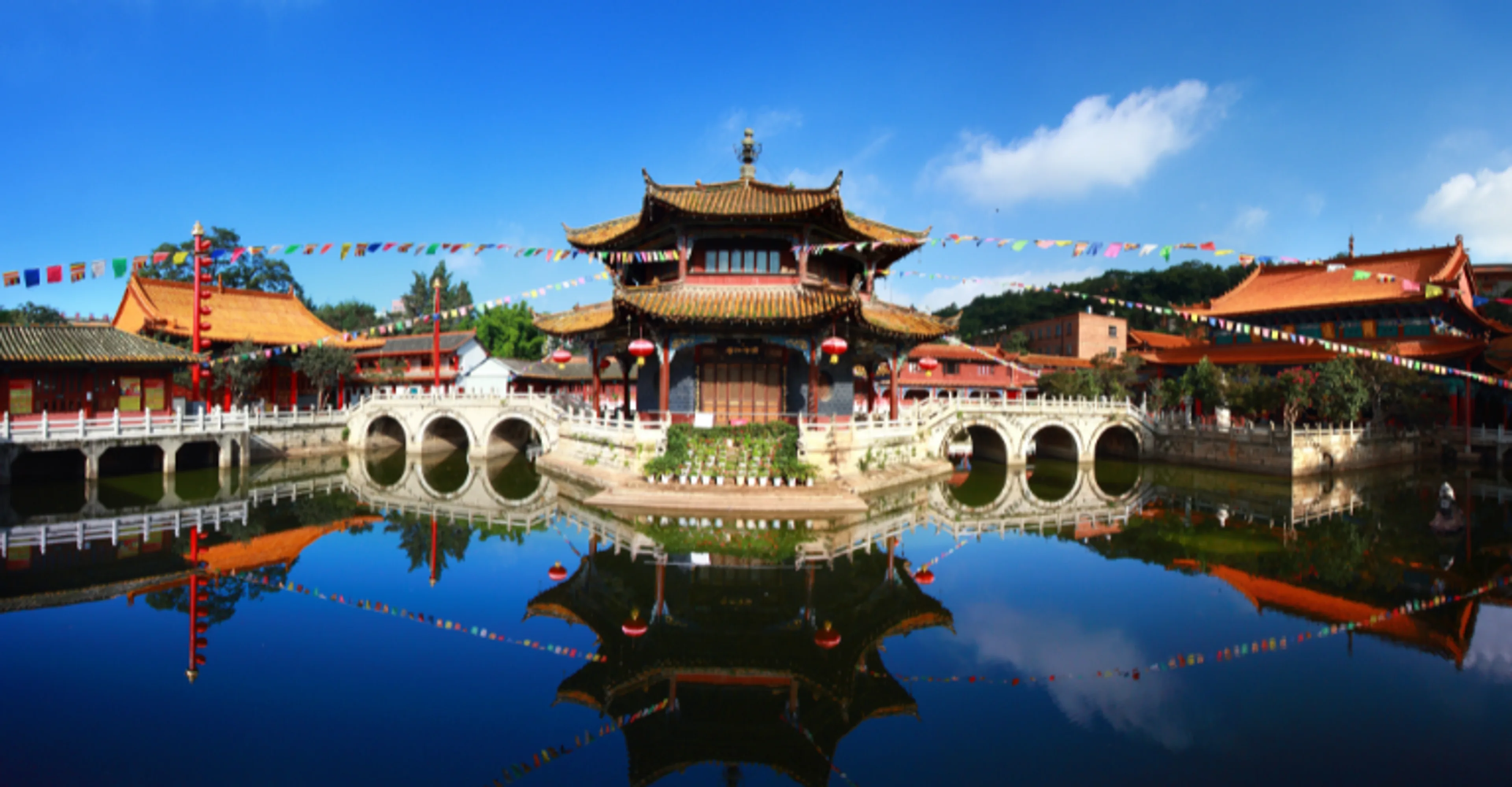 A traditional Chinese temple with an ornate curved roof sits at the center of a pond, connected by white stone arched bridges, with colorful prayer flags strung overhead and the scene mirrored in the calm water under a blue sky.