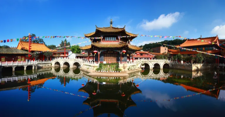 A traditional Chinese temple with an ornate curved roof sits at the center of a pond, connected by white stone arched bridges, with colorful prayer flags strung overhead and the scene mirrored in the calm water under a blue sky.