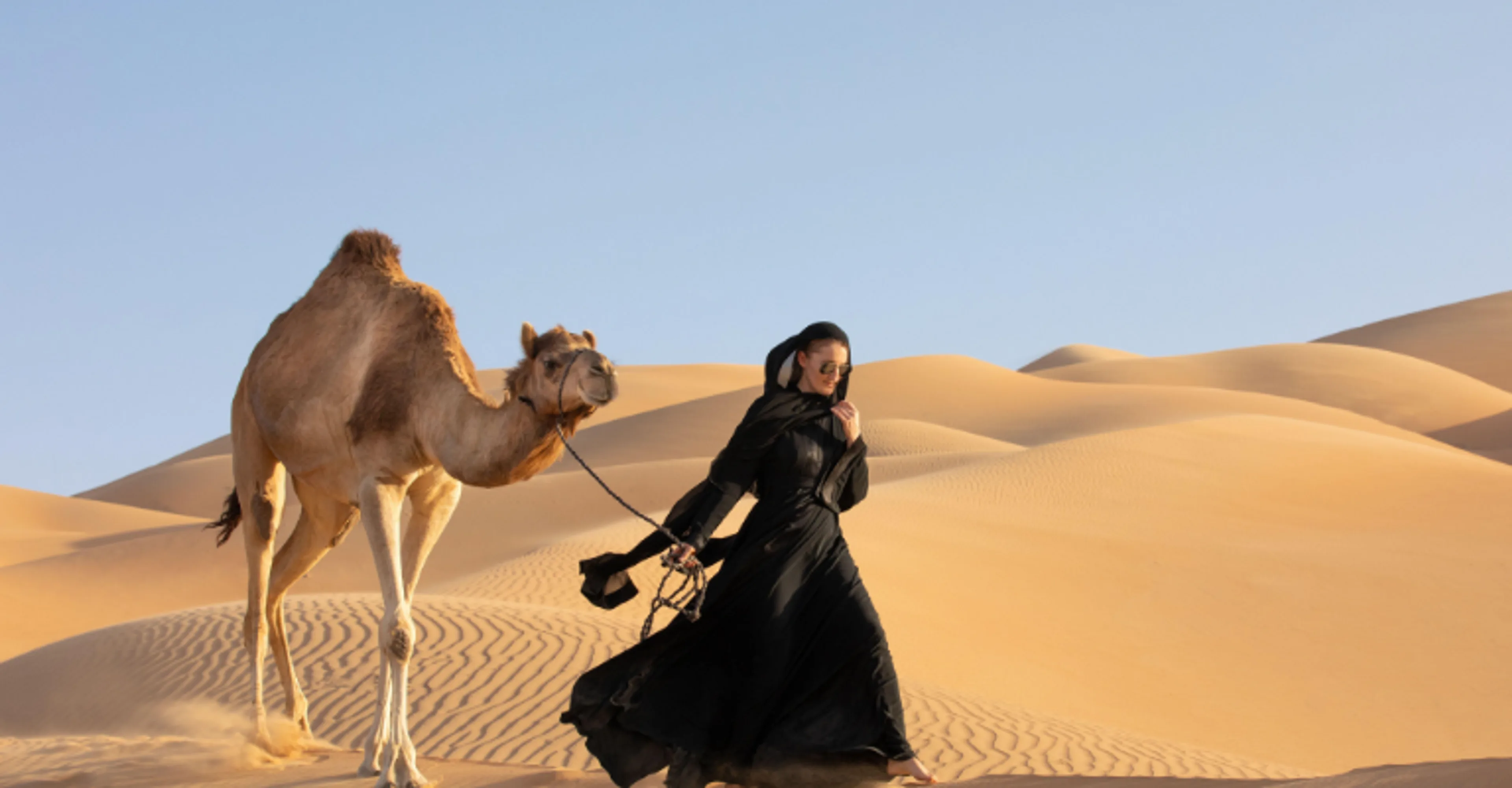 Woman in traditional black attire walking a camel across UAE desert dunes