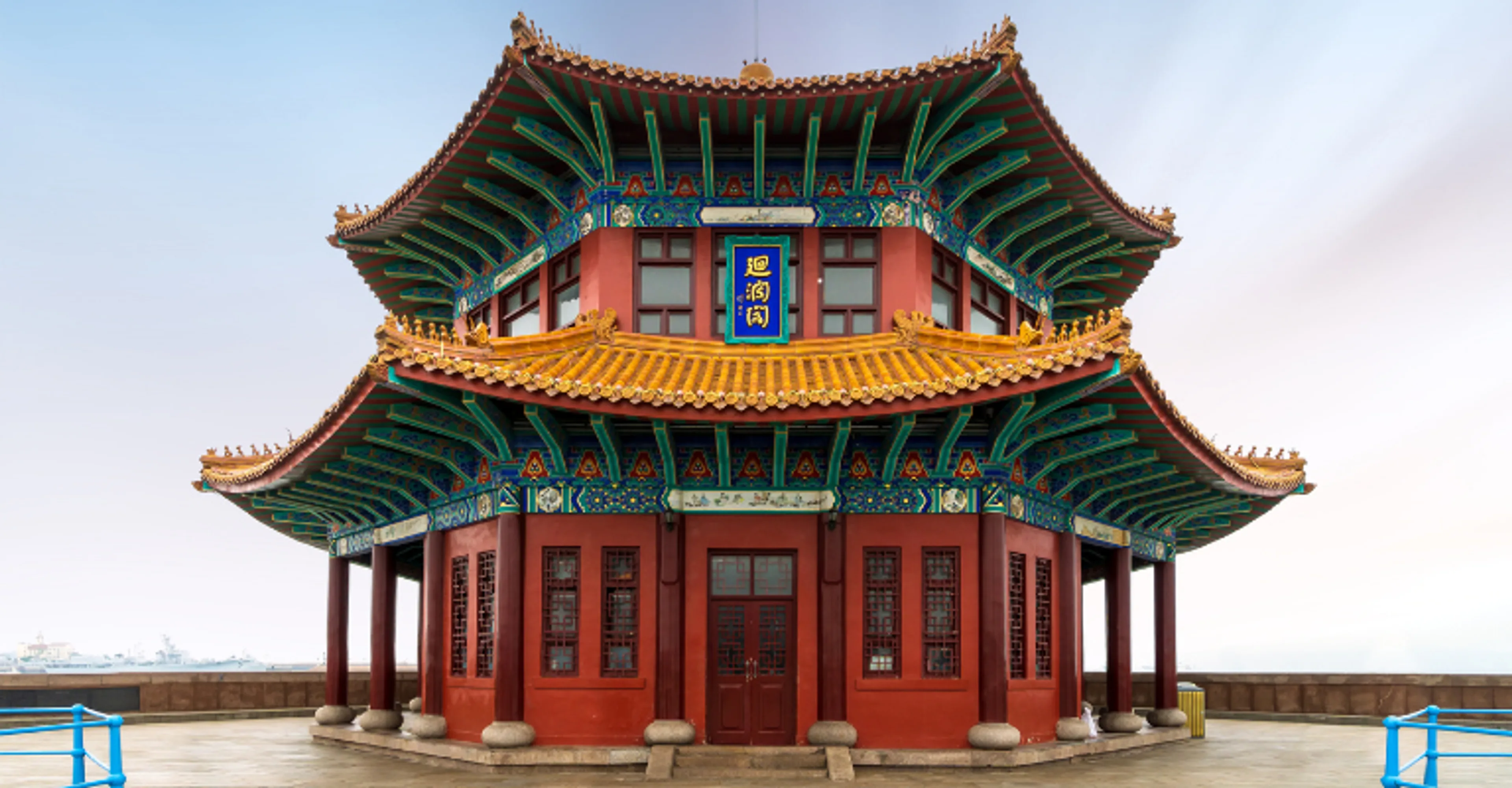 A ornate two-tiered Chinese pavilion with vividly painted red, green, and gold eaves and a blue signboard with gold Chinese characters, set against a pale sky.