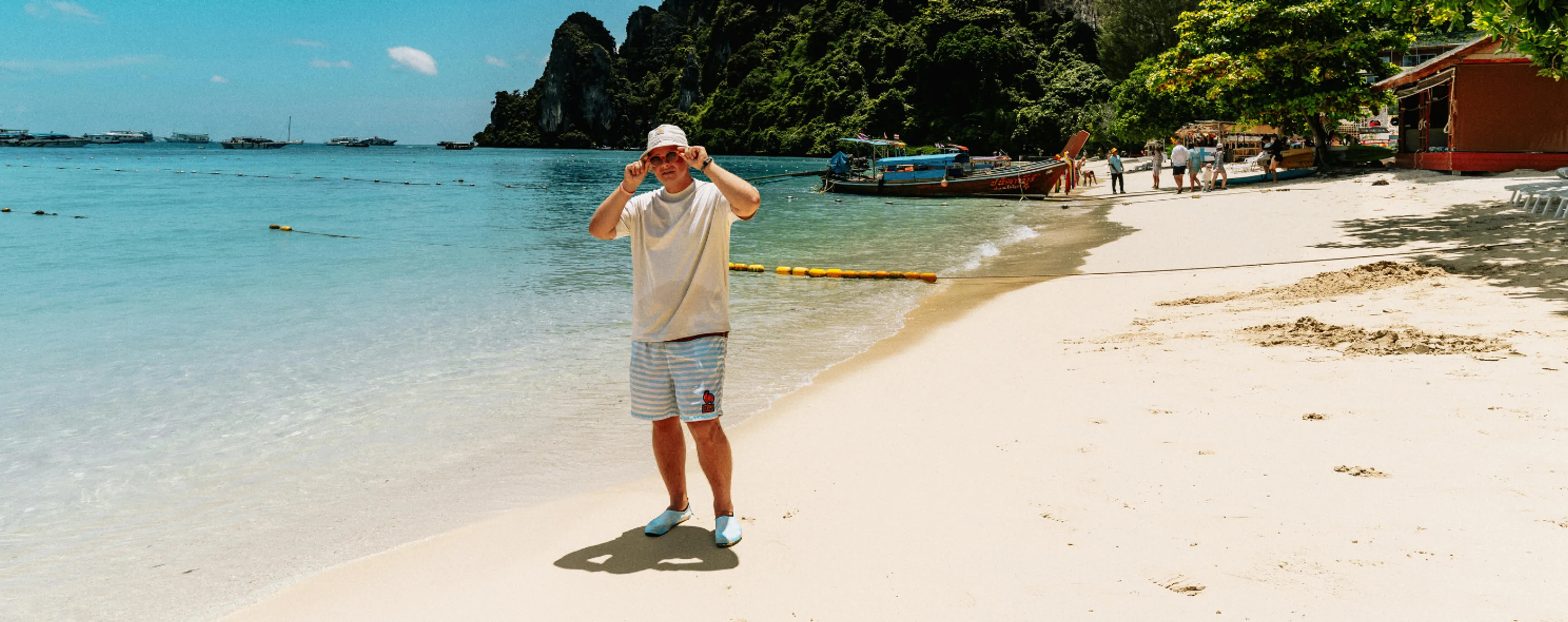 Man in a bucket hat standing on a white sand beach with longtail boats and limestone cliffs in Thailand