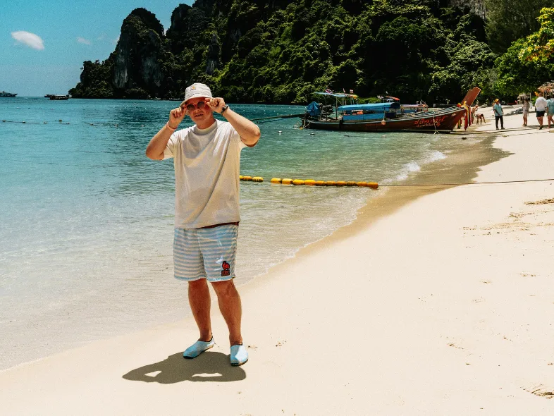 Man in a bucket hat standing on a white sand beach with longtail boats and limestone cliffs in Thailand