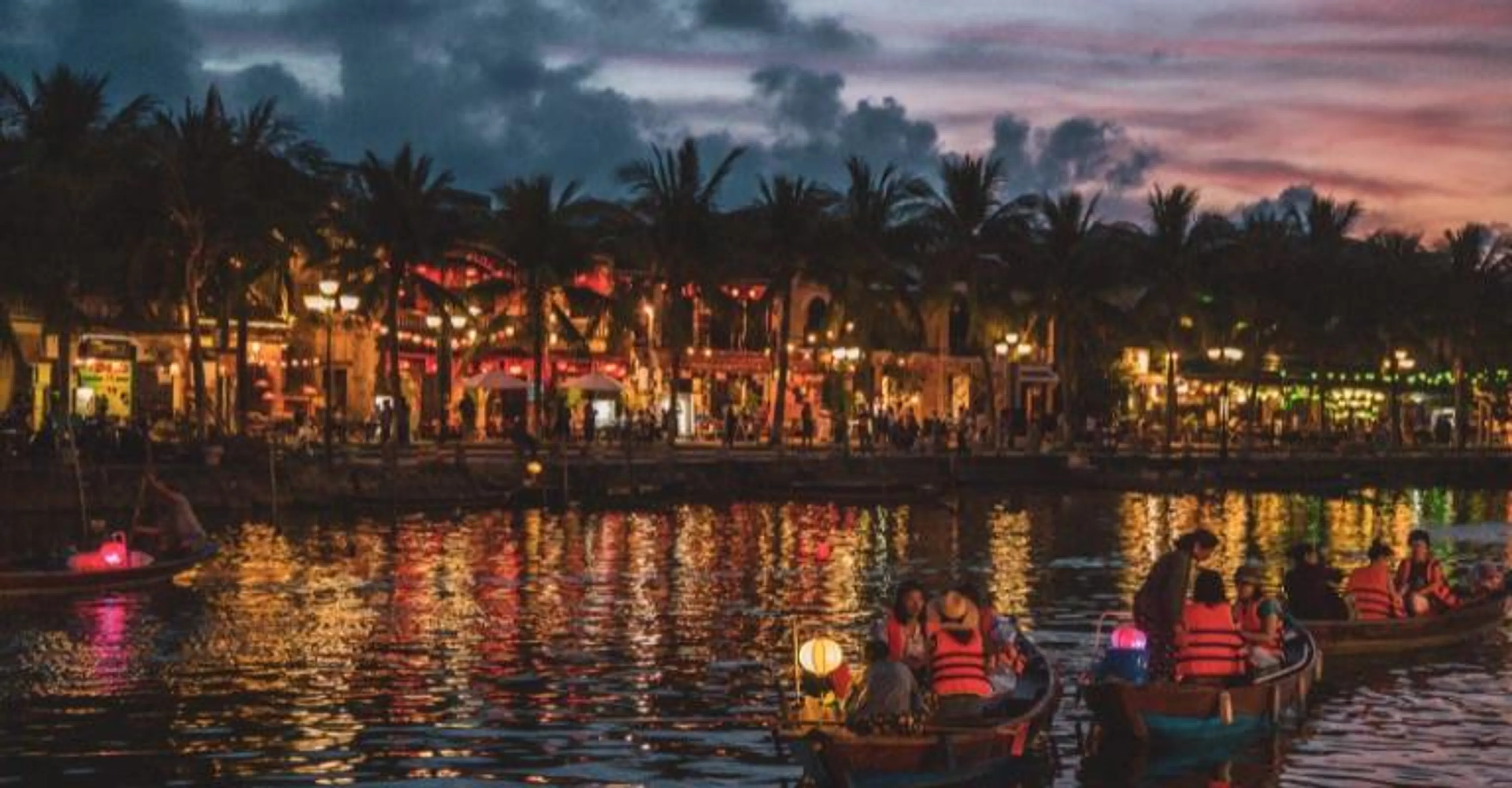 Lantern-lit boats gliding along the river in front of Hoi An's riverside restaurants at night