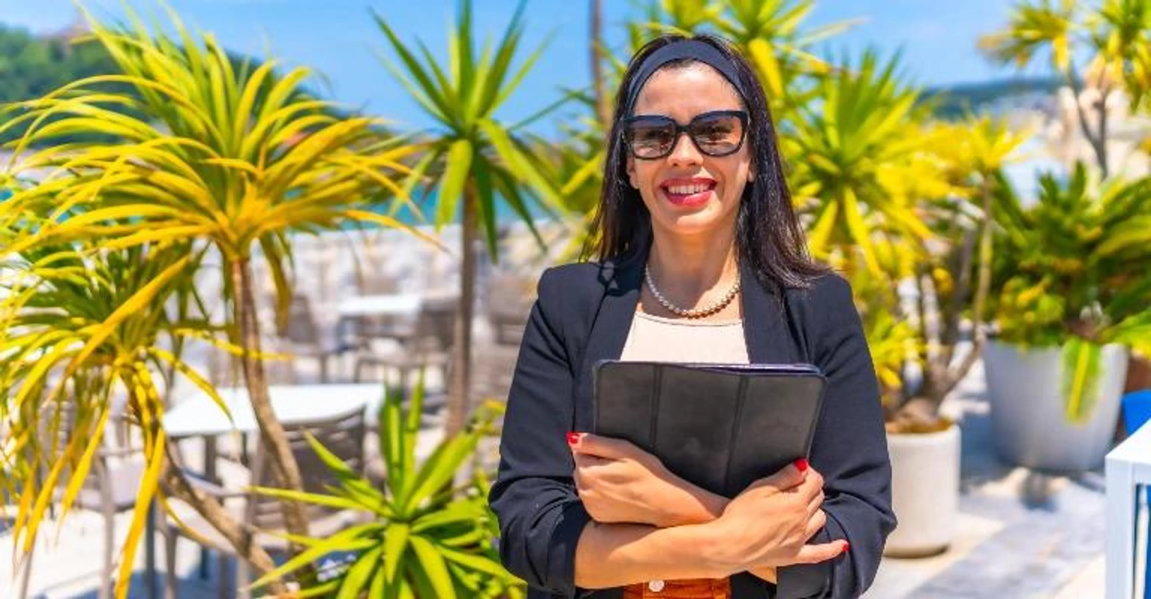 A businesswoman holding a folder, smiling outdoors near palm trees.