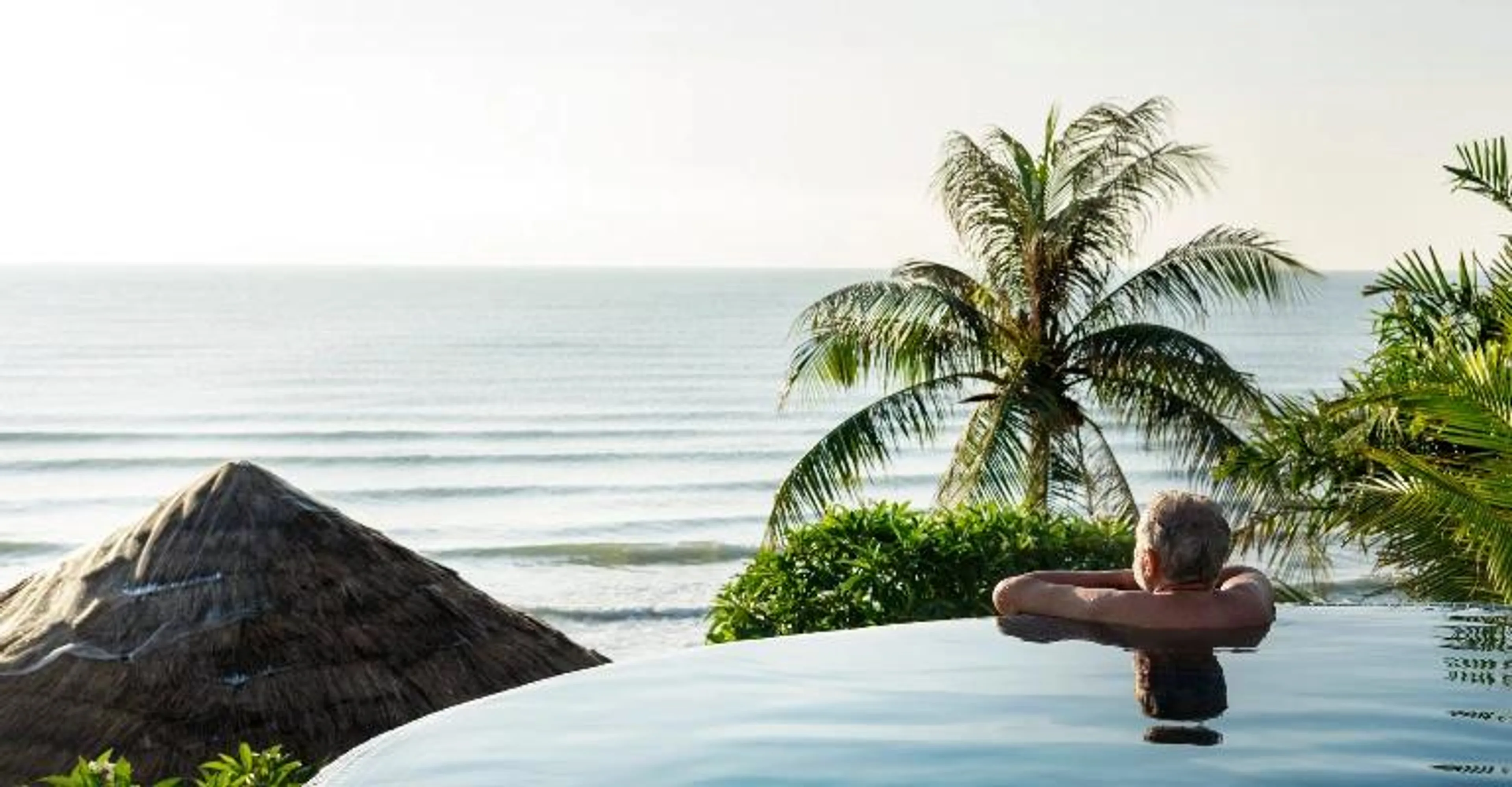 A person relaxes at the edge of an infinity pool overlooking the ocean, resting their arms on the pool's rim as gentle waves roll in under a hazy sky, with a palm tree and thatched-roof structure nearby