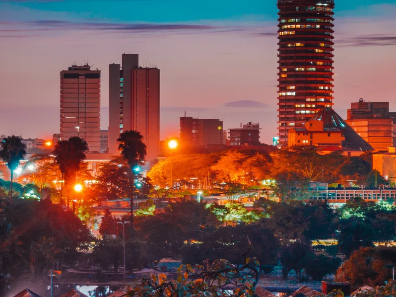 Nairobi's skyline glows at dusk, with illuminated high-rises rising above a foreground of dense city trees