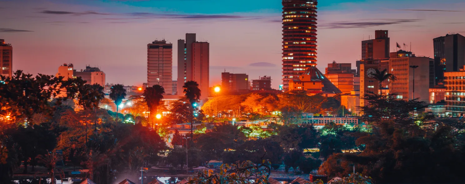 Nairobi's skyline glows at dusk, with illuminated high-rises rising above a foreground of dense city trees