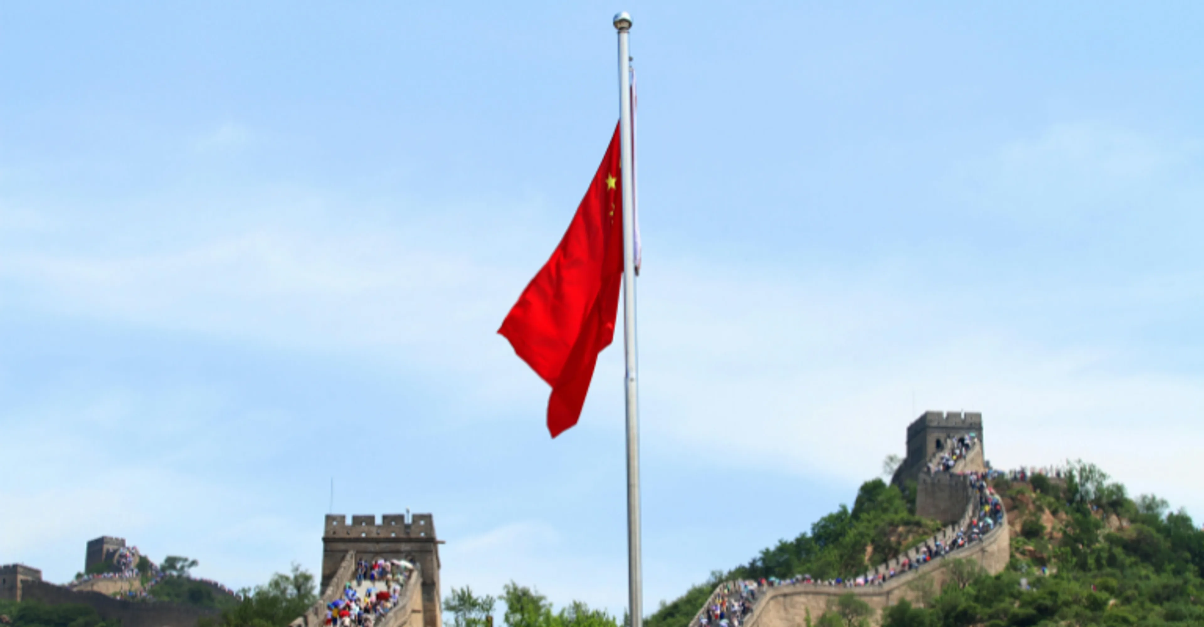 The red flag of China with its yellow stars flies from a pole against a blue sky, with the Great Wall of China winding along forested hills in the background, crowded with visitors