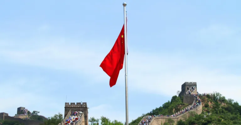 The red flag of China with its yellow stars flies from a pole against a blue sky, with the Great Wall of China winding along forested hills in the background, crowded with visitors