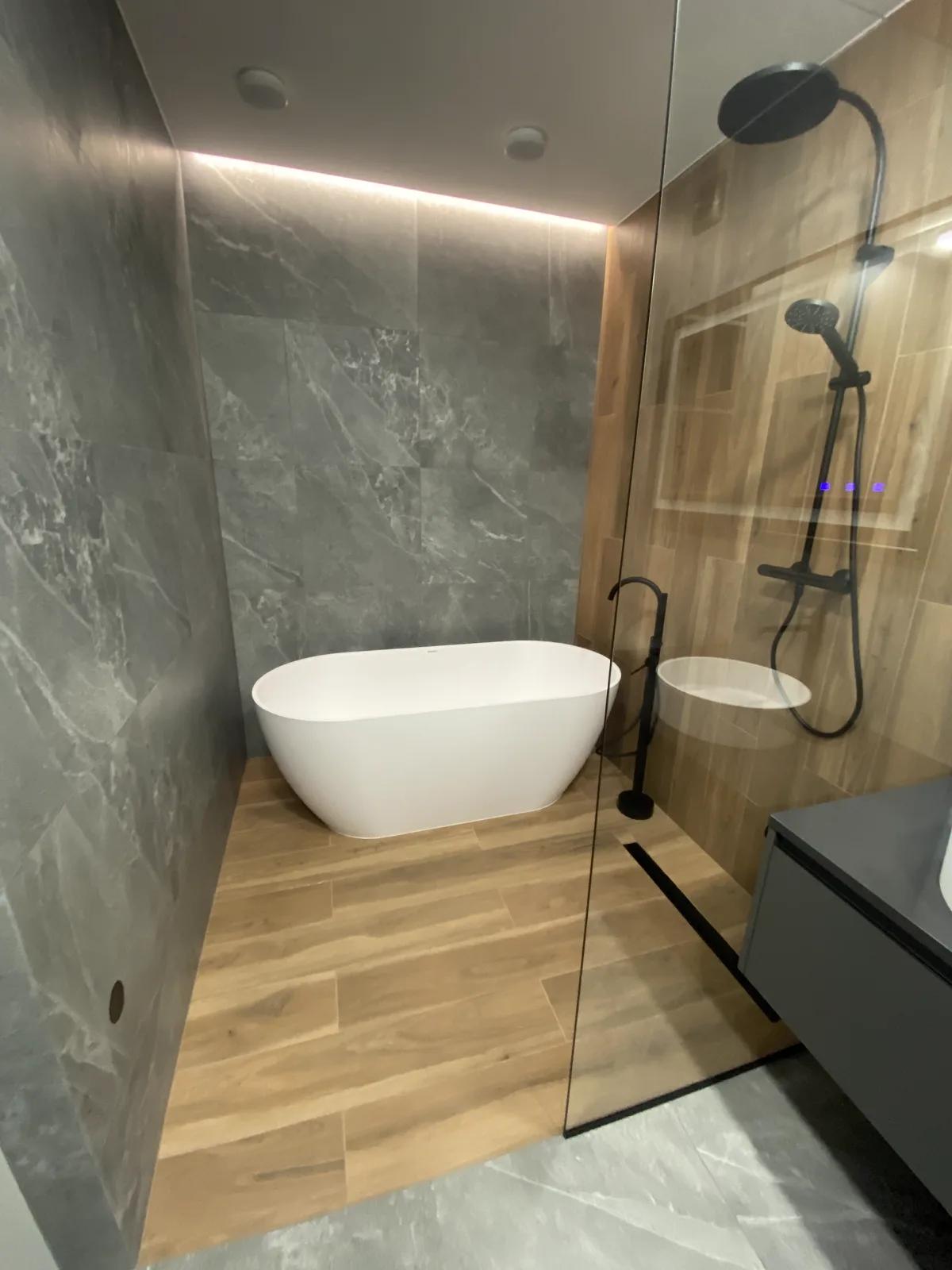 Baño con bañera exenta sobre suelo efecto madera, ducha con mampara y grifería negra