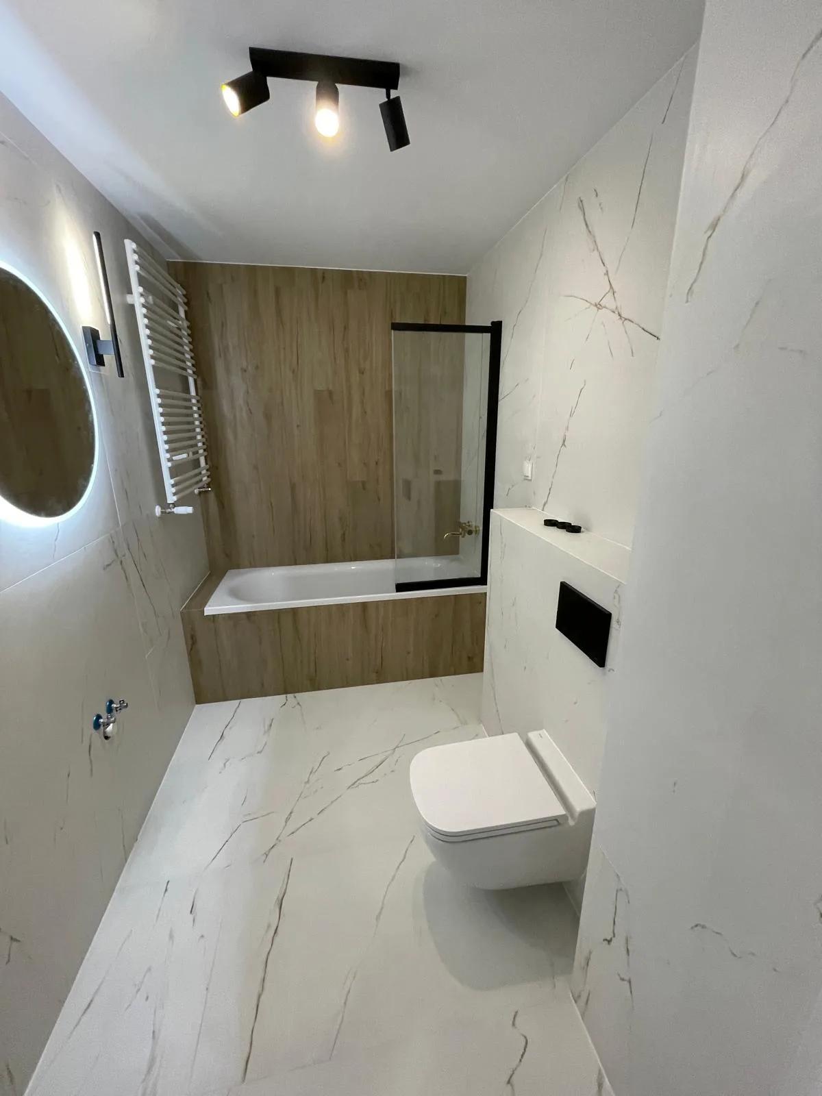 Baño reformado con porcelánico efecto mármol blanco, frente de madera, grifería negra y espejo redondo con luz