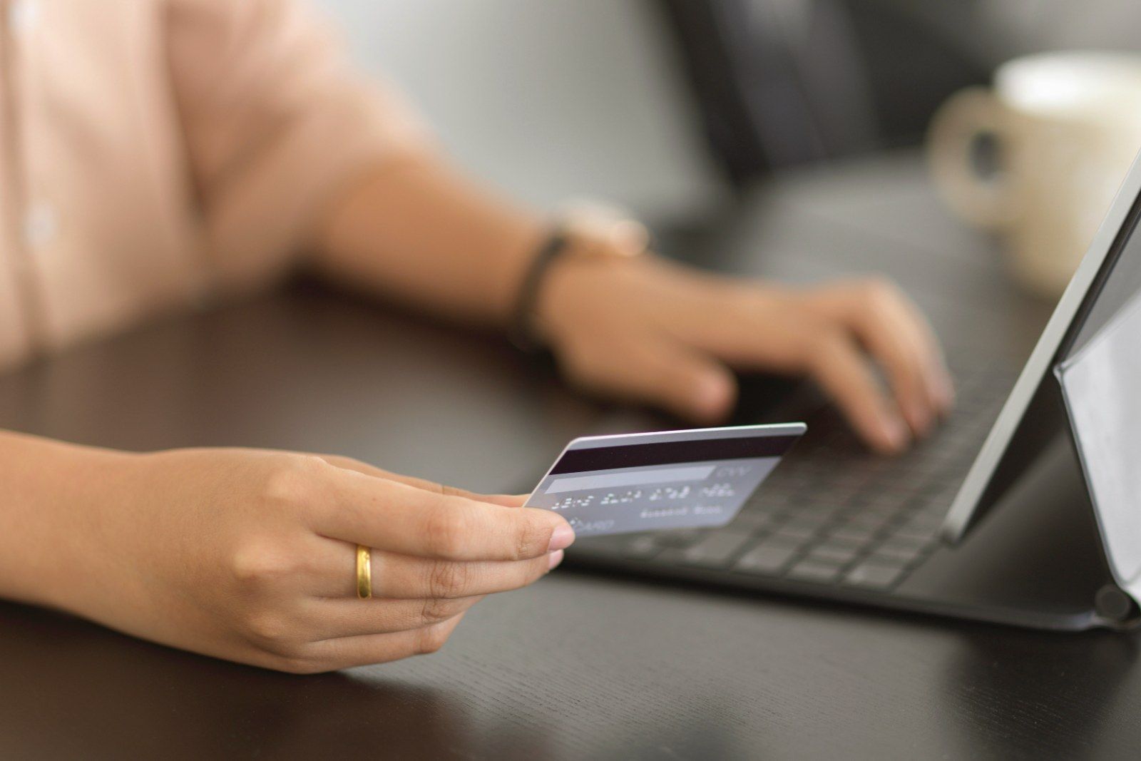 Close view of a person's hands at a laptop, holding a bank card, with a coffee cup behind.