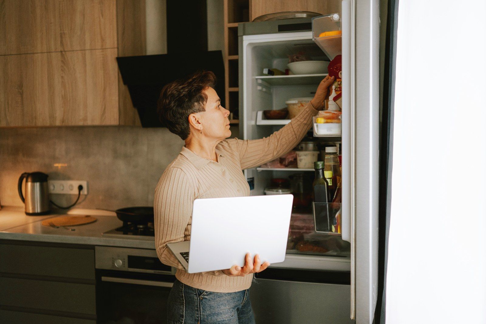 A woman holding a laptop in one hand while reaching into an open fridge.