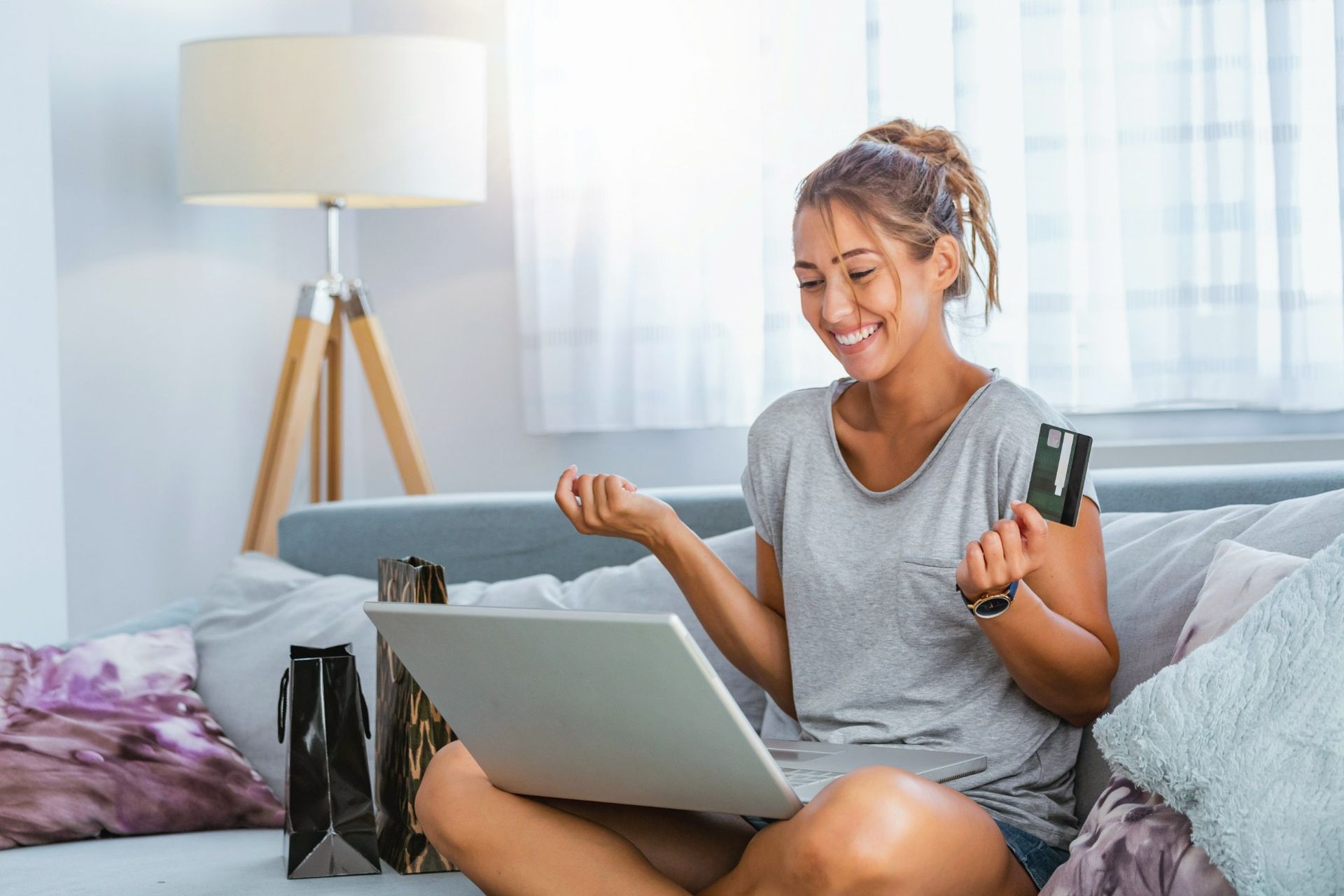 A woman sitting on a bed with a laptop, holding a bank card, with shopping bags beside her.